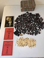 Photo showing Mahjongg tiles spread out with original box and three rule/scoring booklets and scoring sticks on white surface.