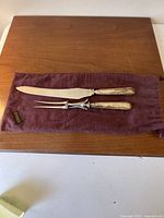 Photo showing carving knife and carving fork on purple felt pouch on wooden surface.