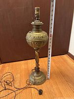 Full view of the brass oil lamp base with measuring tape indicating approximately 23 inches tall, twisted stem and detailed engraving on base and reservoir.