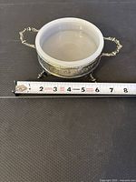 Glass bowl set inside an ornate silver footed holder with handles, viewed top-down with measuring tape for scale showing about 6 inches in diameter.