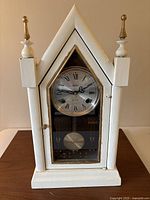Front view of the white hand painted wooden clock with peaked top and gold-tipped finials showing the round clock face with Roman numerals behind a glass door.