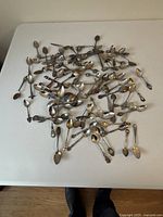 Overview photo showing a large group of vintage and antique collector spoons spread out on a white surface to display quantity and general condition.
