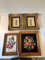 Four framed needlepoint floral artworks grouped against a white wall. Two are in ornate gold frames with flower needlepoints on cream fabric; two are wood-framed with floral needlepoints, one on cream fabric and one on black fabric.
