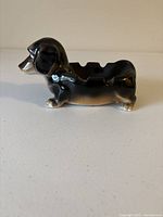Side view of porcelain ashtray shaped like a dachshund dog showing glossy dark brown and black coloring.