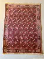 Full view of the miniature carpet showing floral and geometric border patterns in dark pink and beige tones with cream fringe.