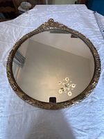 Full view of the round mirror showing the gold gilded frame with ornate floral and leaf motifs and clear reflective glass.