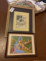 Two framed paintings stacked, one with a river and boats scene, other with a detailed group scene
