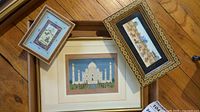 Three framed Indian and Asian art prints on wooden floor. One depicts the Taj Mahal, the others are traditional scenes with riders and camels.
