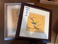 Two framed artworks stacked leaning: front is a yellow background with green nightingale bird and calligraphy in a dark wood frame; back is a floral butterfly themed print in a silver frame.