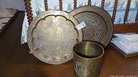 Two brass engraved charger style plates/platter alongside cylindrical brass canister on wooden floor.
