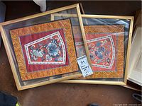 Photo showing both framed embroidered textiles stacked together, displaying overall size and framing.