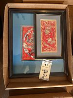 Photo of two framed Asian textile needlepoint artworks featuring dragon motifs on red fabric in black frames and blue mats, shown inside a cardboard box.