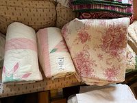 Photo showing three folded bed linens on a patterned sofa: two quilts with pink and white patchwork, and one folded comforter/bed skirt with floral pink and white design.