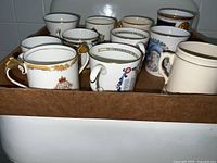 Photo of 11 bone china mugs with various designs in a cardboard box