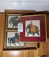 Three framed art prints, two with horse themes and one with elephant and rider theme, arranged inside a box on wooden floor.