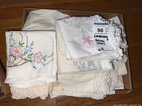 Box of embroidered pillow cases showing multiple folded pillow cases with floral embroidery and lace trims on cream colored fabrics.