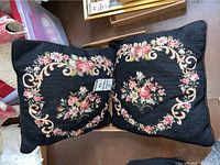 Two black square pillows with floral embroidery on one side placed together to show matching designs.