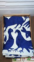 Folded white tablecloth with blue embroidered bird silhouettes visible inside a shallow cardboard box.