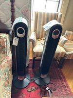 Two black DREO tower fans model DR HTF001 standing on wooden floor next to floral furniture and a striped armchair with sunlight from window