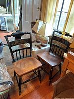 Two Hitchcock style chairs side by side showing seat and backrest details, placed on wooden floor in a room with various household items.