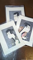 Three Japanese Kabuki caricature art prints stacked, each in plastic sleeve on grey surface