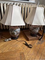 Pair of Asian themed ceramic table lamps with beige fabric shades and cords lying on a wooden floor near a radiator.