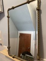 Rectangular mirror with a twisted brass rope frame hanging on a wall, showing the room reflection.