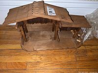 Overall view of wooden nativity manger showing roof, base, and structural supports on a wooden floor.