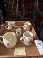 Overview of five Queen Elizabeth II commemorative mugs in a box, showing various portraits, crests, and gold detailing.
