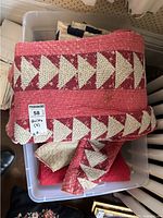 Stack of folded quilts including a prominent red, white, and pink quilt with triangle patterns inside a plastic tote