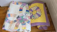 Three folded quilts stacked in corner showing patchwork ring pattern quilt on top and two yellow quilts with purple borders beneath