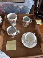 Four pieces of King George V commemorative china including two mugs, a tumbler, and cup with saucer shown in a cardboard box.