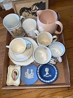 Top view of a wooden tray containing various royal family-themed ceramics including mugs, teacups, a pink pitcher, and Wedgwood commemorative blue plates.