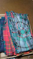 Photo showing four folded Scottish kilts in various tartan patterns in cardboard box on floor