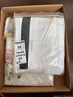 Two shower curtains folded and packaged in a cardboard box, one white with dark vertical stripe, the other floral patterned.