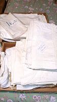 Multiple white linens folded and stacked in cardboard boxes on floral tablecloth.