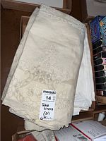 Folded cream-colored embroidered tablecloths with floral and geometric cutwork placed in a box, photographed from above to show details and lot label.