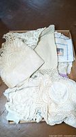 Box filled with folded vintage linens including crocheted doilies, embroidered napkins, and assorted fabric pieces.