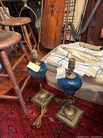 View of two vintage matching blue glass table lamps with ornate metal bases on a red rug near wooden chairs and textiles.