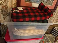 Red and black plaid blanket folded on top of clear plastic storage bin containing other blankets