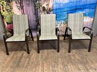 Three stackable aluminum sling chairs with bronze powder-coated finish, arranged side by side on a wooden floor with a beach and floral backdrop.