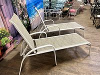Two white aluminum frame chaise lounge chairs with beige fabric slings and adjustable backs, shown side by side on a wooden floor.
