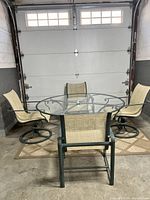 Full view of round glass top table and four chairs arranged around it, showing verdigris finish and sling fabric seating.