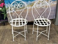 Front view of two white metal bistro chairs with lattice seats and sweetheart back scroll designs, showing off-white powder coated finish.