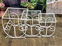 Full set of three white iron nesting tables with decorative scrollwork and glass tops shown nested together.