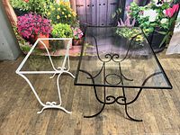 Photo shows both black and white wrought iron side tables with glass tops, placed on a wooden floor with a floral backdrop.