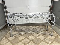 Front and left side view of vintage wrought iron console table base showing overall shape and scroll design, with no tabletop attached, placed on tiled surface indoors.