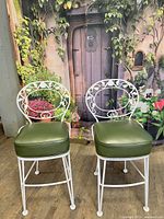 Two Woodard Chantilly Rose pattern metal barstools with white powder-coated finish and original green vinyl cushions, positioned in front of floral backdrop.