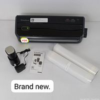 Top view of FoodSaver main vacuum sealer unit, two sealing rolls, portable vacuum pump with charging cable, and user manual arranged on white surface. Label on vacuum sealer states brand new.