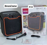 Full view of ION Sport XL speaker next to its original box with the wired microphone, power cable, and manual laid out in front.